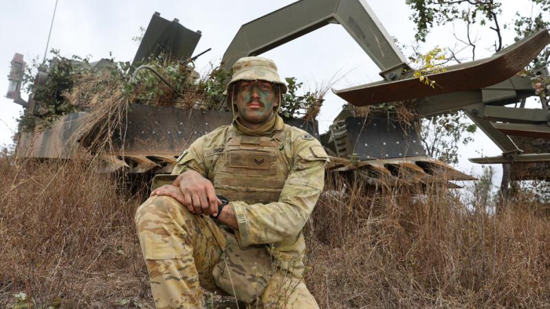 A man in military fatigues crouches slightly down a hill from a large military vehicle.