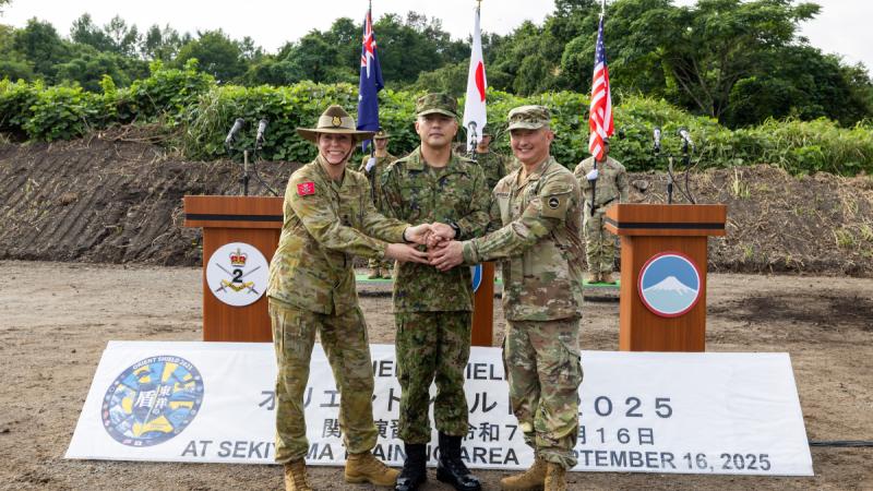 Three people in uniform shaking hands on a parade ground, standing in front of lecterns and the flags of Australia, Japan and the United States.