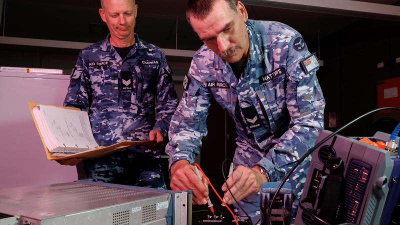 Two men in Air Force uniforms, one holding a ring binder, the other testing some electronic equipment.
