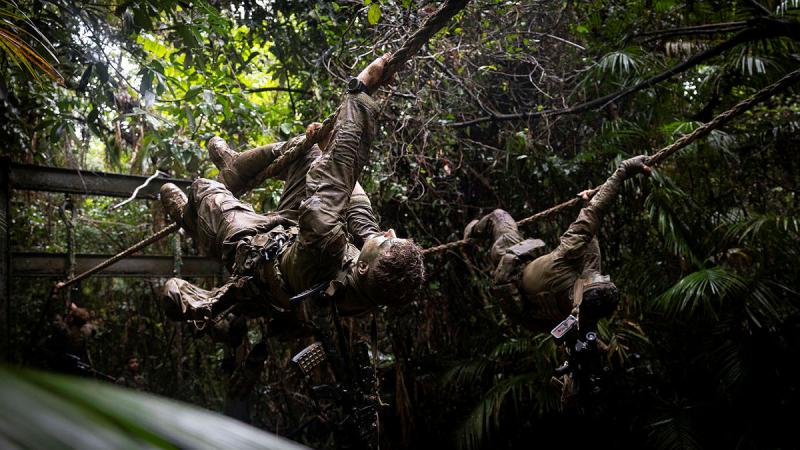 Two soldiers are upside down on a rope in a wet and muddy jungle.