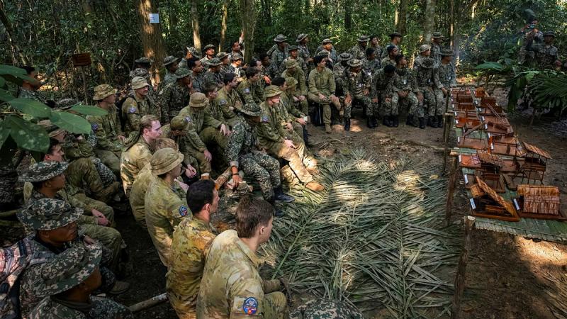 Military personnel sit in a large group with jungle craft demonstrations in front of them.