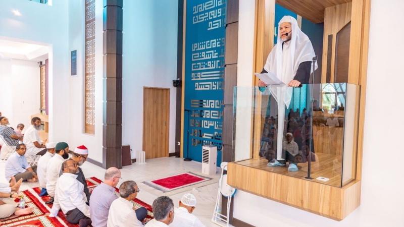An Navy Imam in Islamic robes speaks at a mosque attended by men in civilian clothes. 