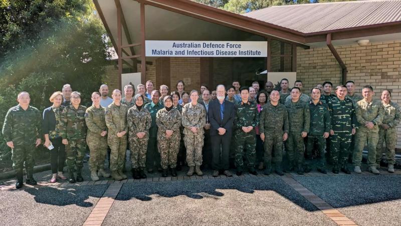 A group photo of approximately 30 people, many in military fatigues, standing outside the Australian Defence Force Malaria and Infectious Disease Institute building.