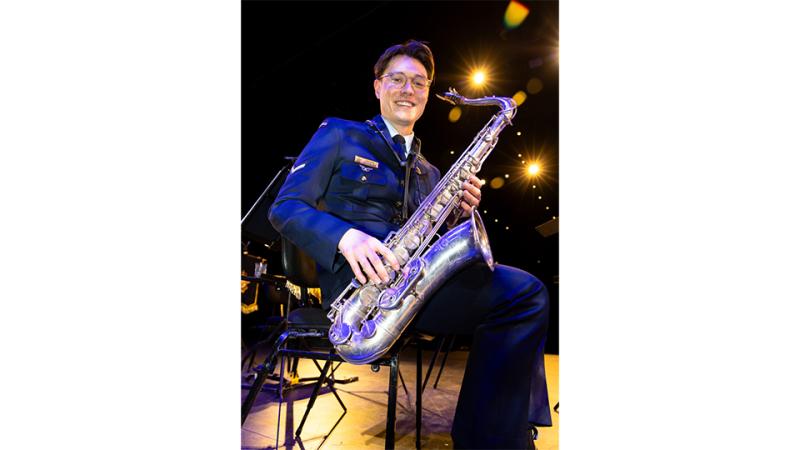 A man in military band uniform sitting on a stage holding a tenor saxophone.