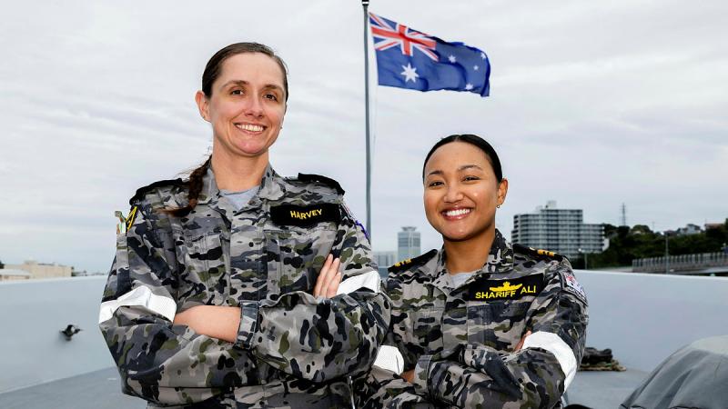 Two female Navy personnel in uniform stand smiling, with their arms crossed, in front of an Australian flag.