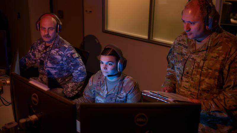 Three uniformed people wearing headsets look at computer screens in a dimly lit room.