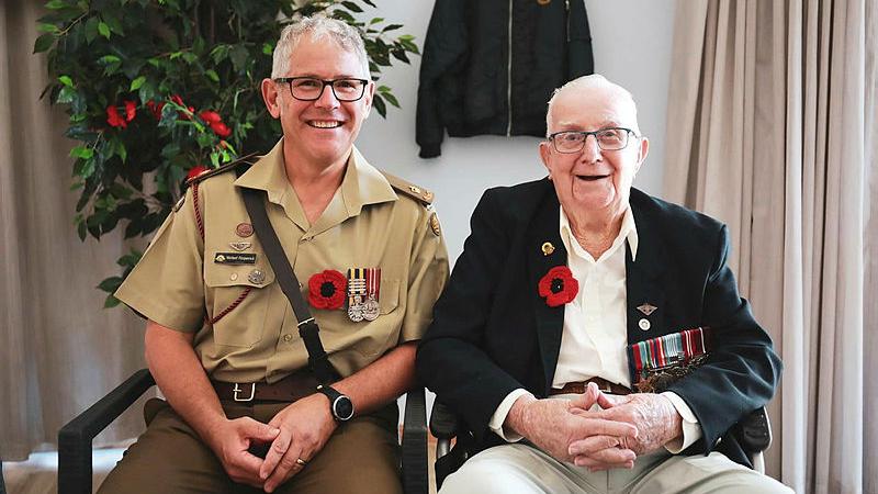 A man in Army uniform sits with another elderly man in civilian clothing, both wearing poppies.