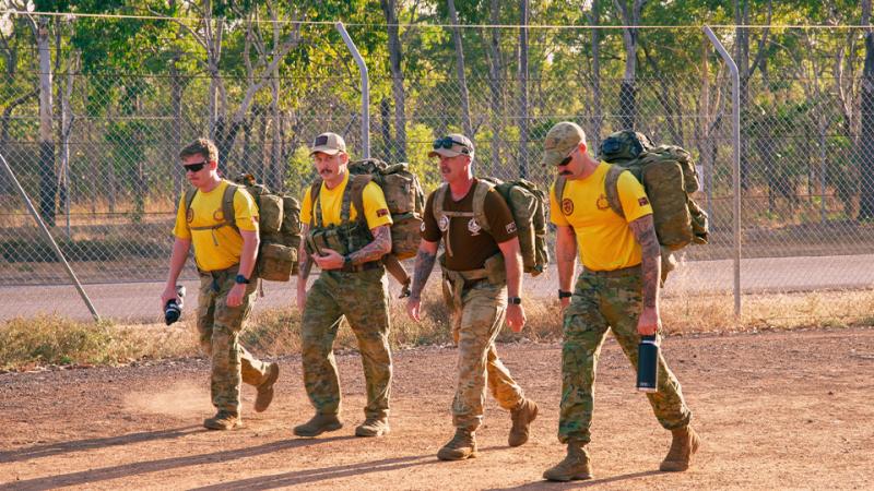Four soldiers wearing heavy packs walk along a dirt track next to a security fence.