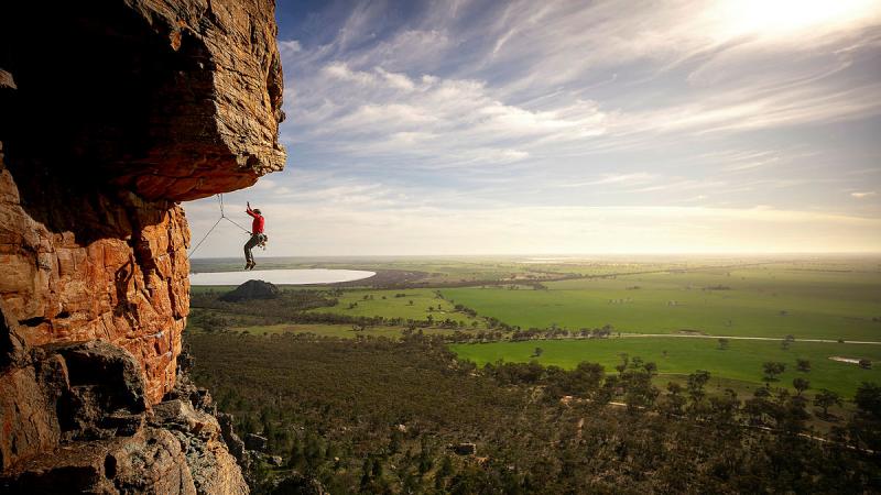 A person hangs on a rope from a rock face with blue sky, sun and green plains in the background.
