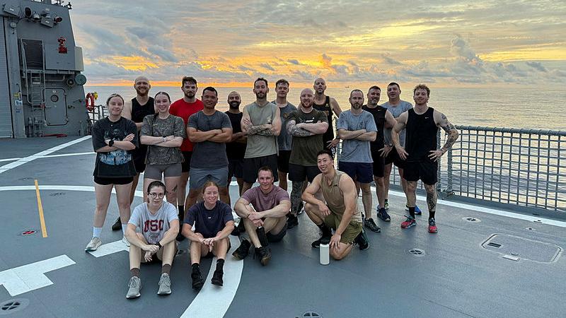 A group of Navy personnel in exercise gear stand and sit in a group on the deck of a ship with a sunrise in the background.