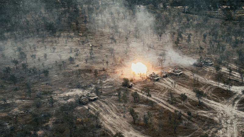 An aerial shot of a group of tanks, one firing, in a bush setting.