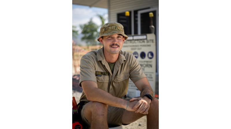 A young man with a moustache sitting outside a worksite.