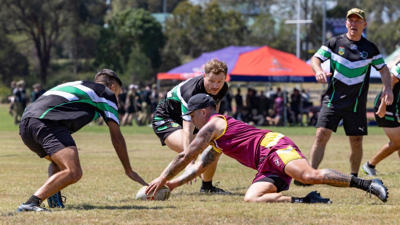 A player in maroon singlet and shorts scores a try by diving between defenders dressed in black with teal-and-white stripes.