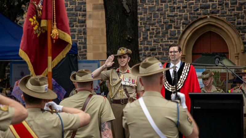 Soldiers stand facing the Chief of Army who is saluting and standing next to a man in a red gown. with a