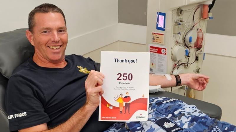 An Air Force aviator in blue camouflage uniform donates blood, lying on a bed next to a machine, holding a 250th donation  certificate.