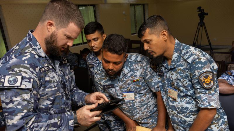 Three men in Bangladesh Air Force uniform look at something a man in Royal Australian Air Force uniform is showing them on a tablet.