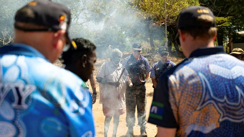 First Nations people and sailors stand in front of a fire with smoke rising.