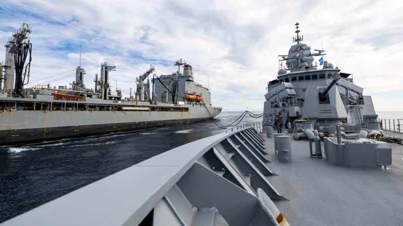 Two military ships sail next to each other, connected by fuel lines.