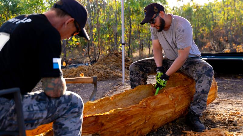 Two Navy personnel use hand tools to carve a wooden canoe.