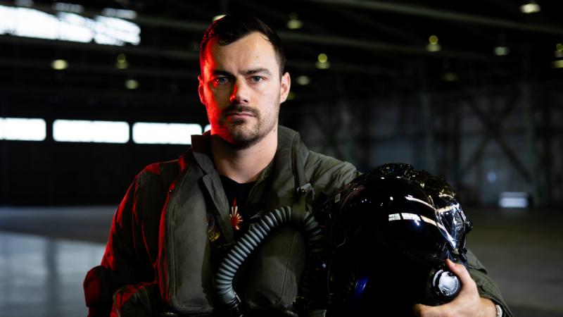 A RAAF pilot stands in a hangar, helmet under his arm, with a red light illuminating one side of his face.