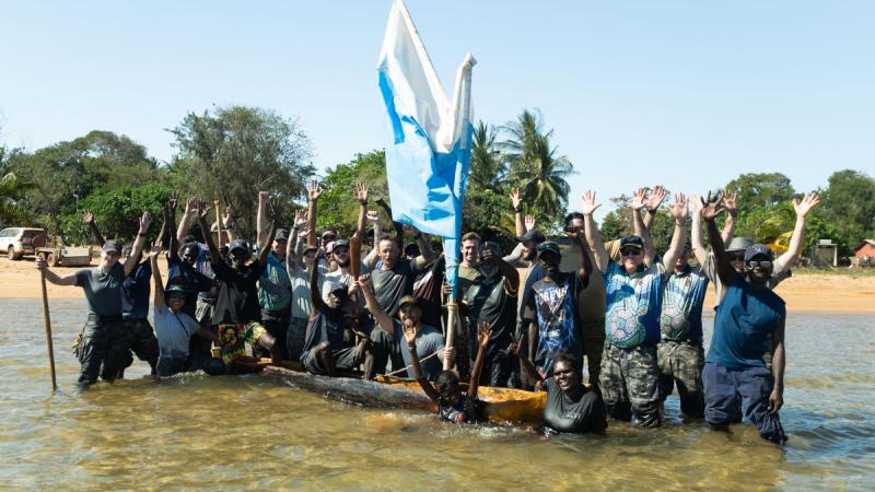 A group of people standing in knee-high water with their arms raised, surrounding a lippa-lippa canoe.