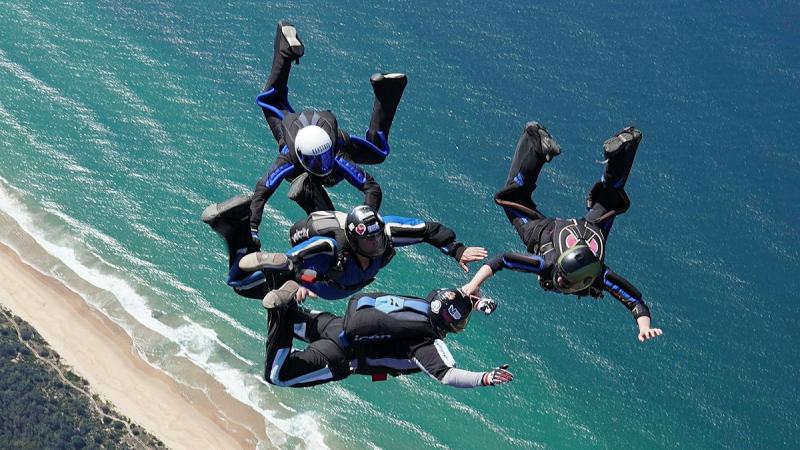 Four people create a formation in mid air above a beach while skydiving.