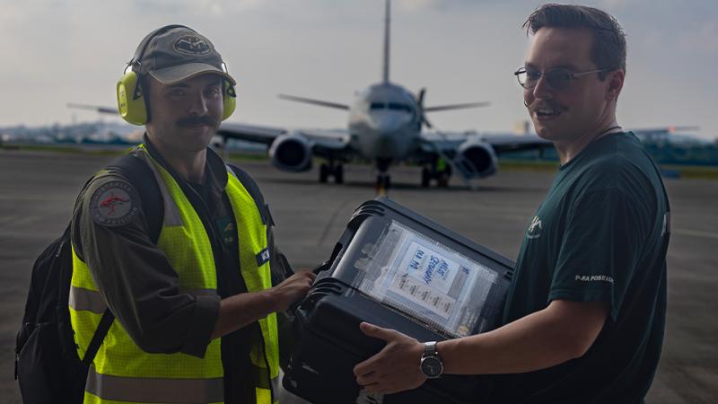 Two men exchanging a large plastic box while standing on a tarmac with a P-8A Poseidon in the background.