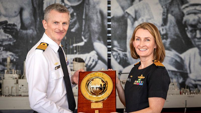 A male Navy officer in a white uniform receives a plaque from a female Navy officer in a blue uniform.