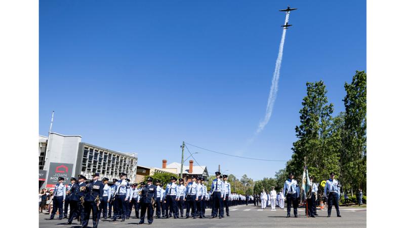 Two small planes fly overhead, contrails behind them, as Air Force personnel march on a city street.