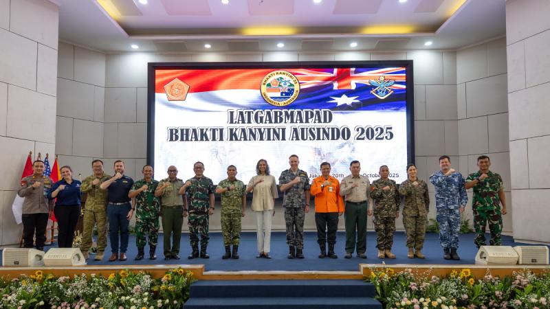 A row of people stand on a stage with fists held in front of their chests, in front of a large screen displaying Indonesian text.