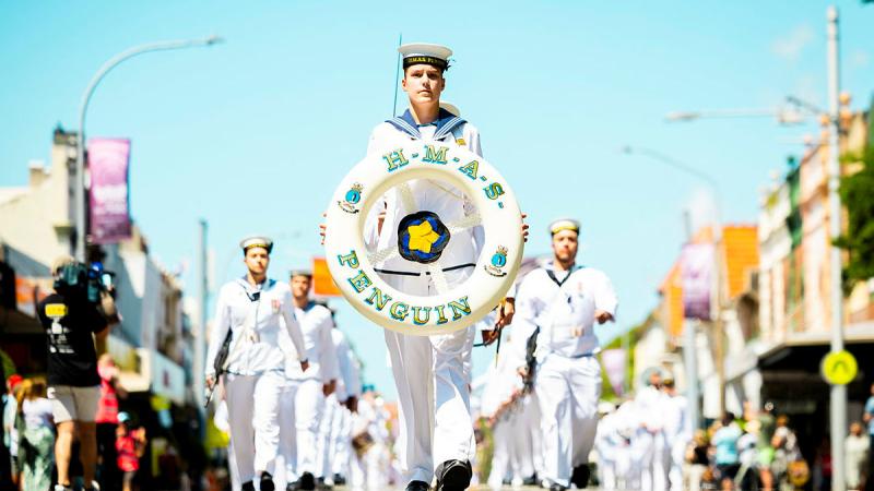 A sailor in white carries a circular lifebuoy with HMAS Penguin printed on it. He marches out the front of other Navy personnel in white uniforms, a crowd looks on.