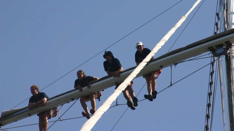 Four crew members on the yard arm of a sailing ship, with blue sky background.