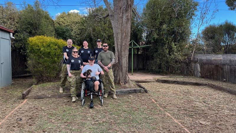 : A man in a wheelchair holding a small dog sits front and centre in a group photo with five other people, taken in a backyard with freshly cut grass.