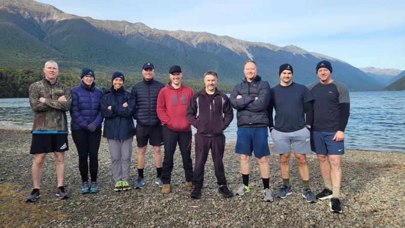 Nine people in outdoor gear stand on a pebble beach with their backs to a lake, with mountains rising across the water.