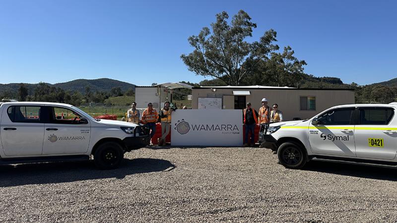 Construction workers stand either side of a business sign, in between two cars at a construction site.