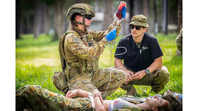 A medic in Army uniform provides blood to a simulated casualty at the point of injury while another soldier observes. 