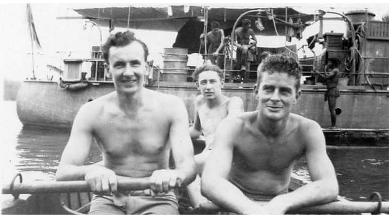 A black and white photo of three shirtless men in a row boat near a larger boat.