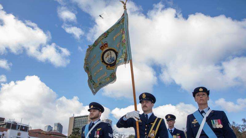 With a helicopter flying high overhead, three people in formal RAAF uniform march in a row in a parade ground, the centre person bearing the Squadron Standard.