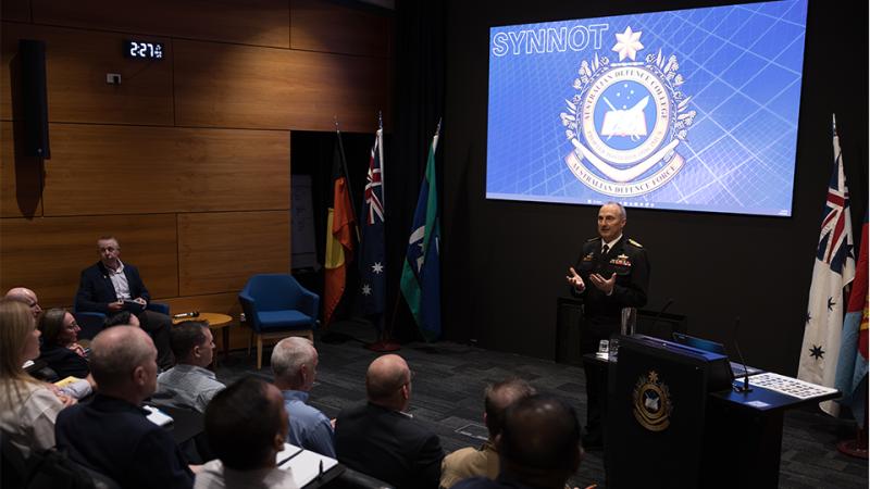 Chief of the Defence Force Admiral David Johnston stands at the front of a lecture theatre giving a talk to an audience, with a presentation displayed on a large screen behind him.