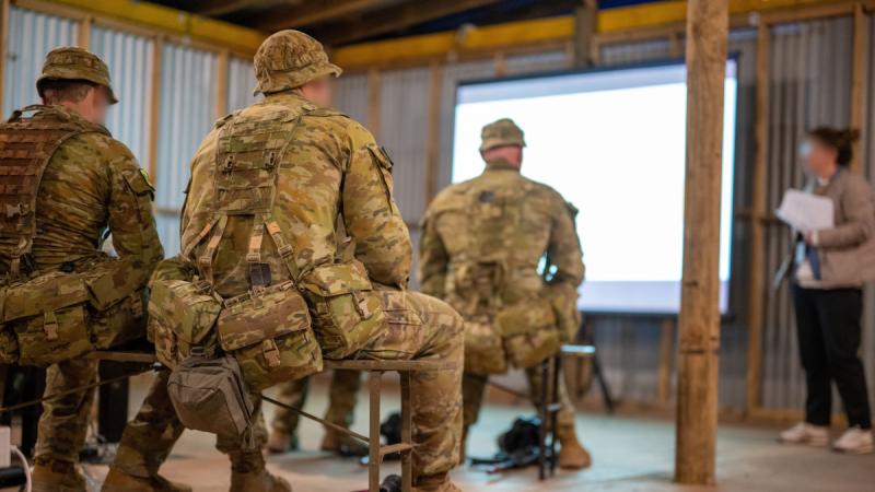 Soldiers, their faces blurred, sit on benches inside a corrugated iron structure, listening to a woman in casual clothes deliver a presentation.