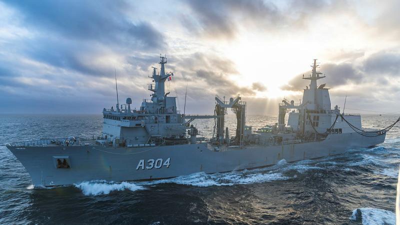 An Australian Navy ship cuts through the waves at sea with a mix of sun and clouds in the sky.