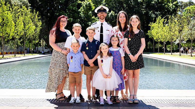 A group of children stand with a Navy officer in uniform in front of a pond.