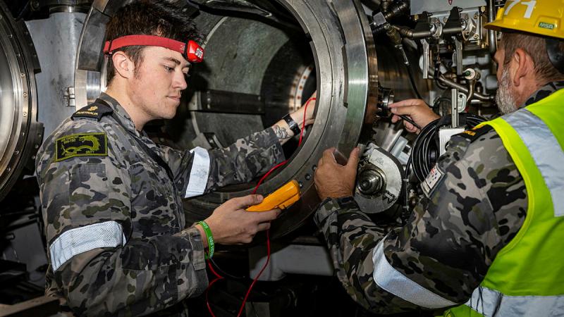 A man looks at a monitoring device he is holding while his other hand touches a round cylinder. Another Navy member assists.