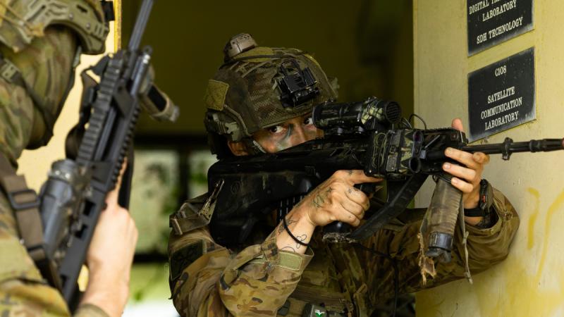 A soldier indoors in full fatigues holds her up weapon and looks through the sight. Another soldier stands beside her, also holding a weapon.