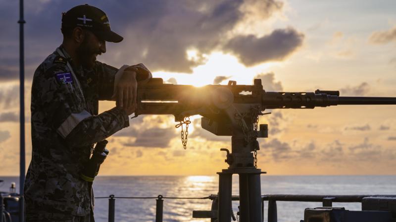A sailor mans a gun on board ship as the sun sets over the ocean.