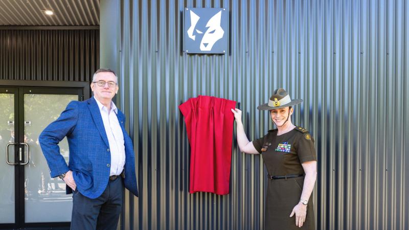 A woman in Army uniform stands with her hand on a small red curtain covering a plaque, with a man dressed in business casual standing on the other side of the plaque.