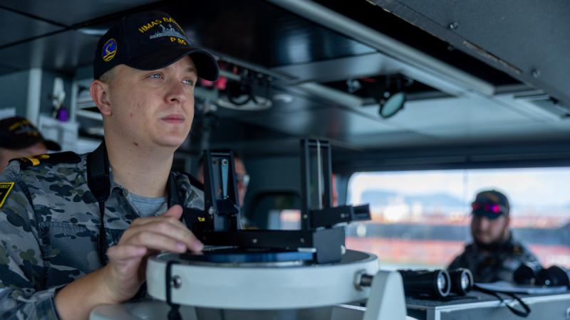 A Navy officer in uniform stands on the bridge, hands resting on a console, looking ahead.