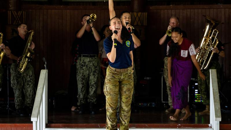 A woman stands on a large speaker in front of a stage, singing into a handheld microphone as a brass band plays behind her.