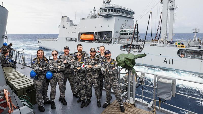 A group of sailors stand on the deck of a ship with another in the background.