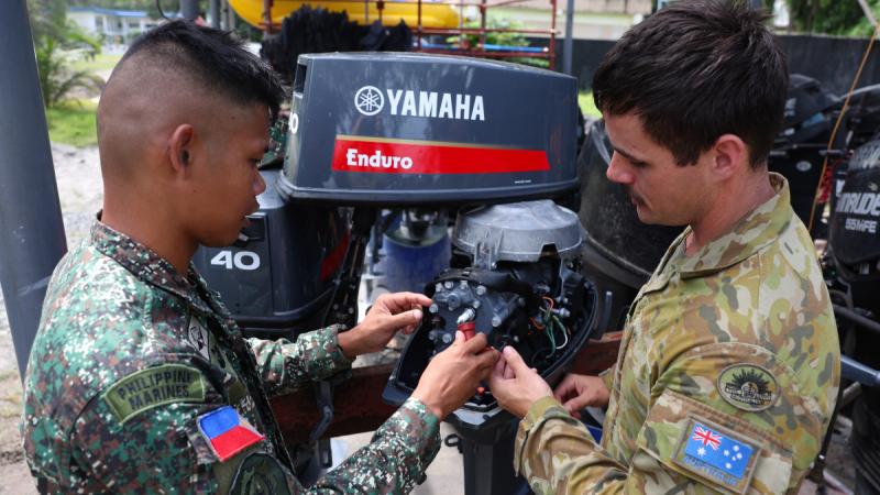 Two men in military uniforms working on an outboard motor together.